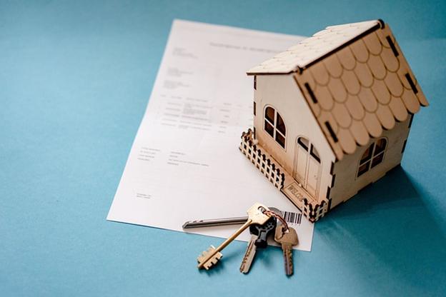 **Alt text:** A small wooden model house and a set of keys are placed on top of a document against a blue background.