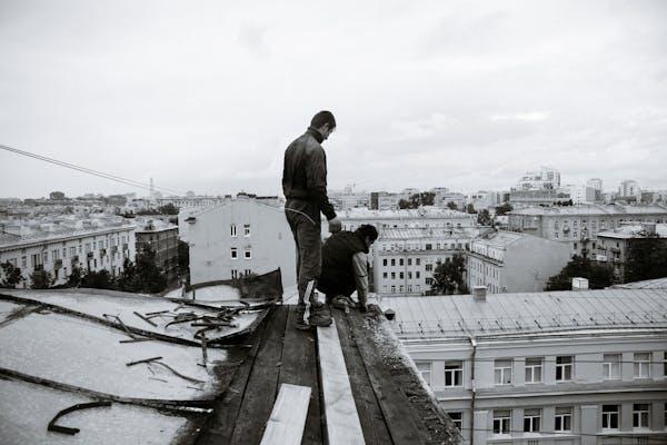 Two people stand on a wooden platform on a rooftop overlooking a cityscape, with one person kneeling and the other standing near the edge.