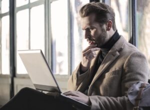 A man in a brown coat sitting by a window, looking intently at a laptop screen with his hand on his chin.