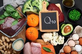 A variety of foods rich in Coenzyme Q10, including broccoli, liver, oranges, avocado, peas, cauliflower, nuts, seeds, salmon, and eggs, arranged around a small chalkboard with "Q10" written on it.