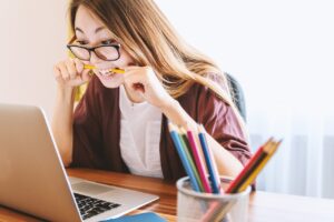 **Alt text:** A woman wearing glasses bites a pencil while looking at a laptop screen, appearing stressed, with a container of colored pencils on the desk.