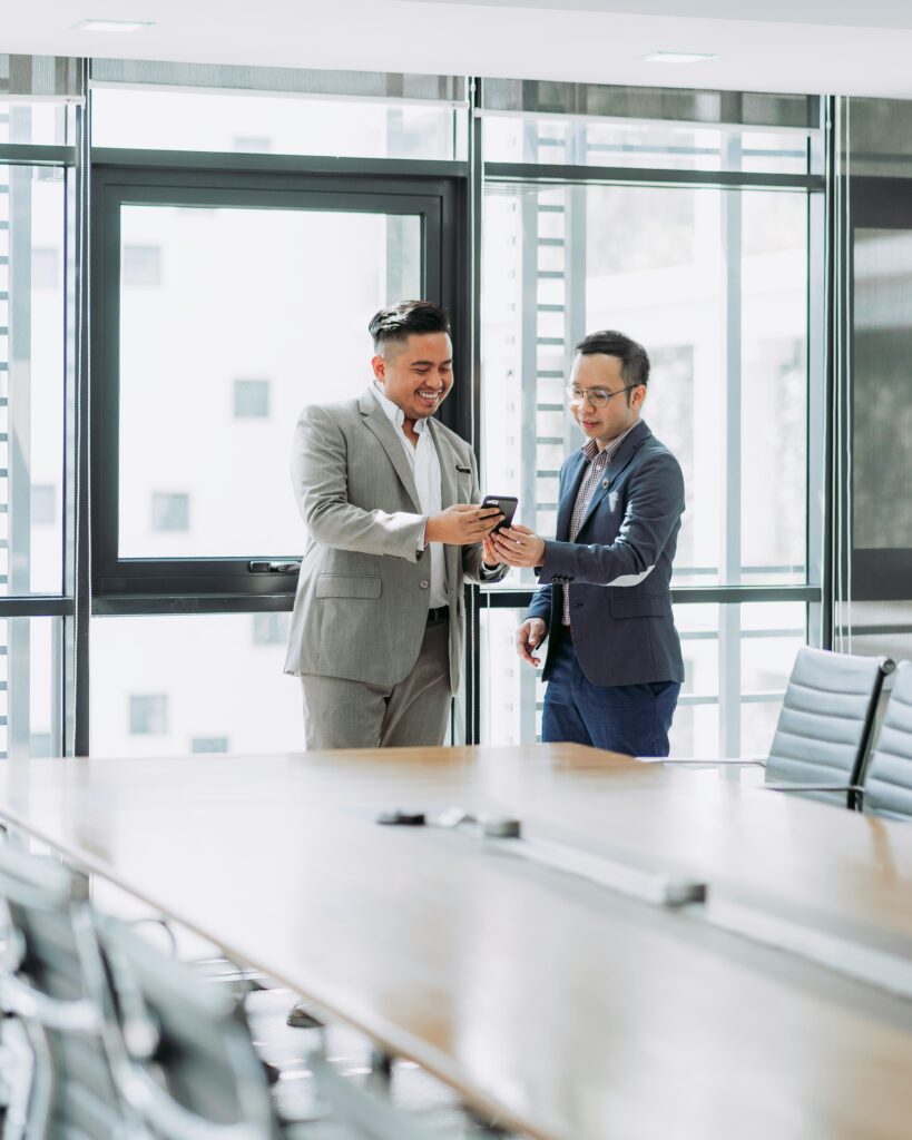 Two men in suits stand in a conference room, smiling and looking at a smartphone.