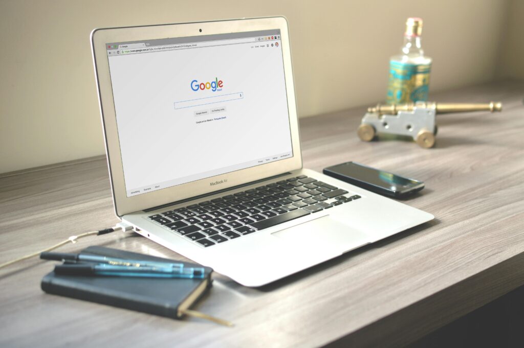 **Alt text:** A laptop displaying the Google homepage is on a wooden desk alongside a notebook with pens, a smartphone, and a decorative miniature cannon.