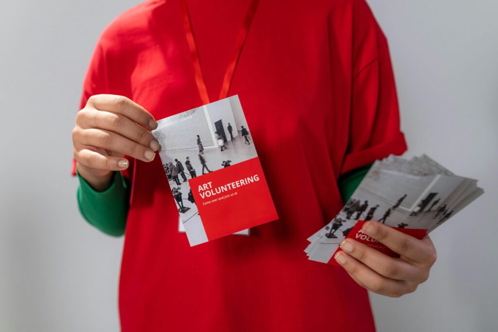 A person in a red shirt holds a stack of flyers titled "Art Volunteering."