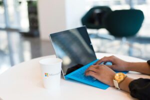 "A person typing on a tablet with a blue keyboard cover next to a white Microsoft-branded paper cup."