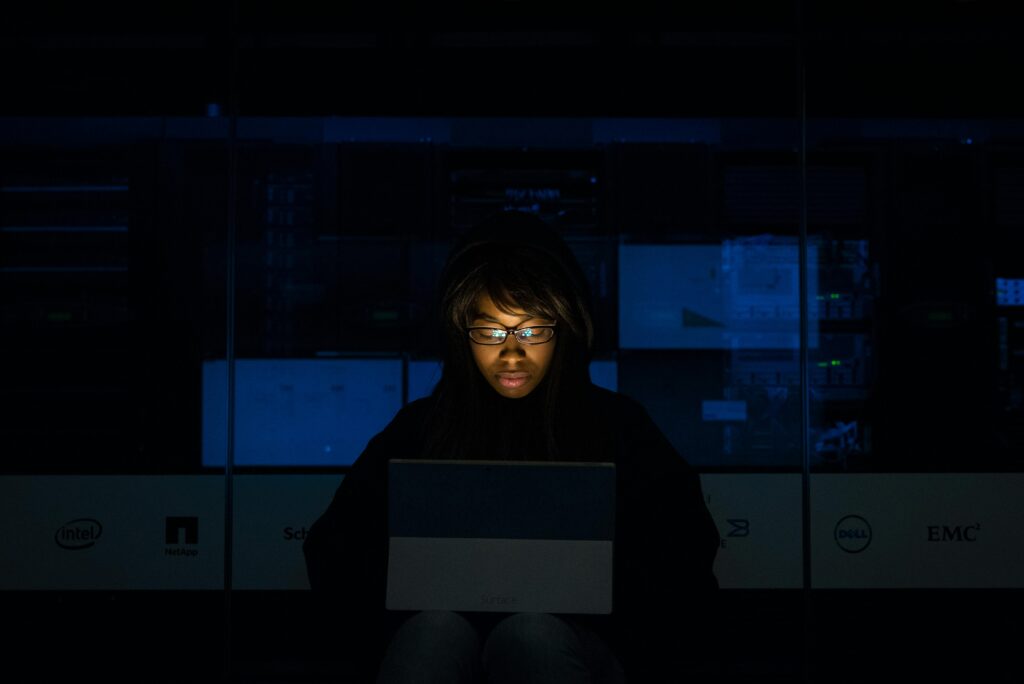 **Alt Text:** A person wearing glasses and a dark hoodie is illuminated by the light of a laptop in a dimly lit room with computer servers in the background.