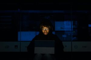 **Alt Text:** A person wearing glasses and a dark hoodie is illuminated by the light of a laptop in a dimly lit room with computer servers in the background.