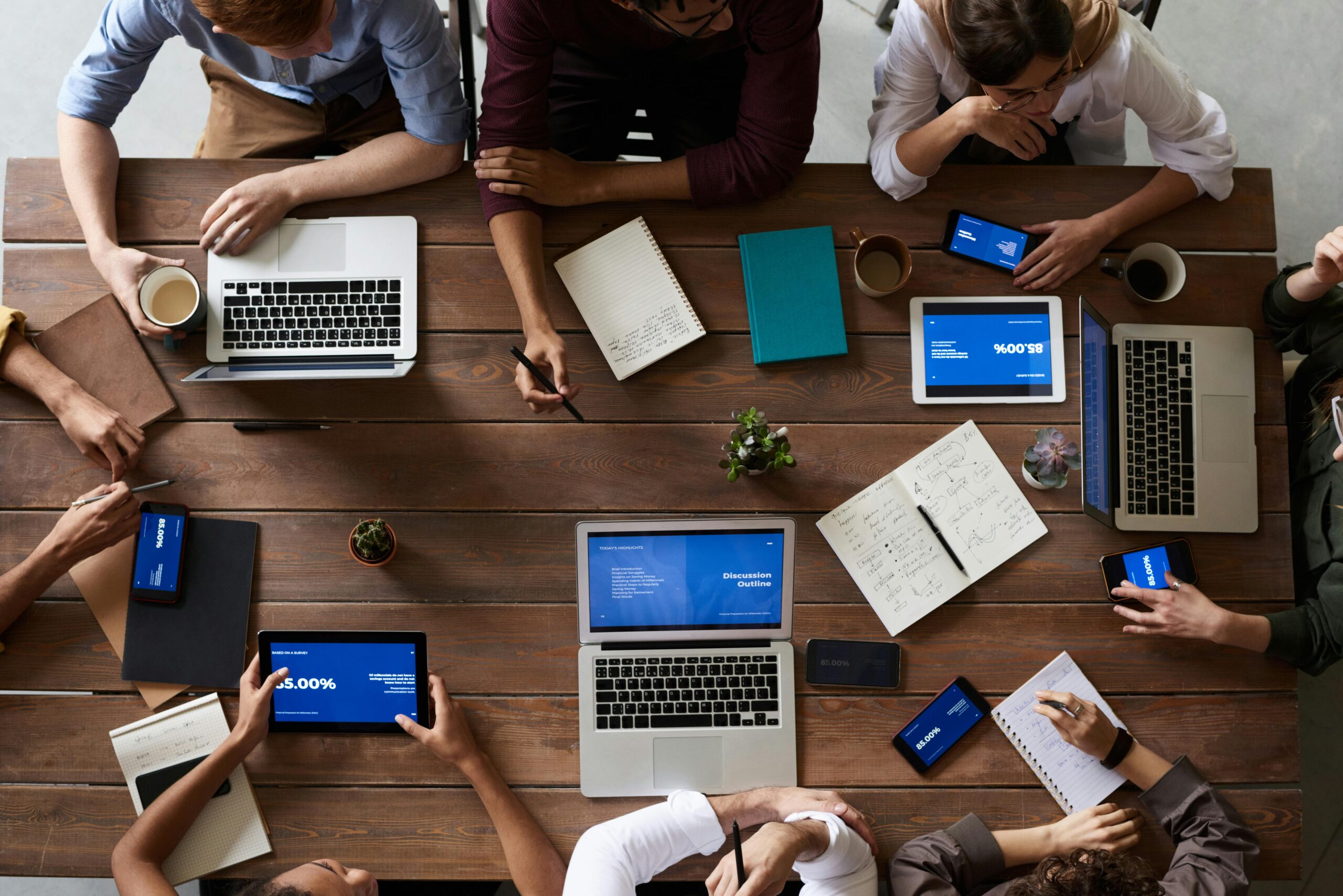 A group of people sitting around a wooden table with laptops, tablets, and notebooks, engaged in a meeting or discussion.