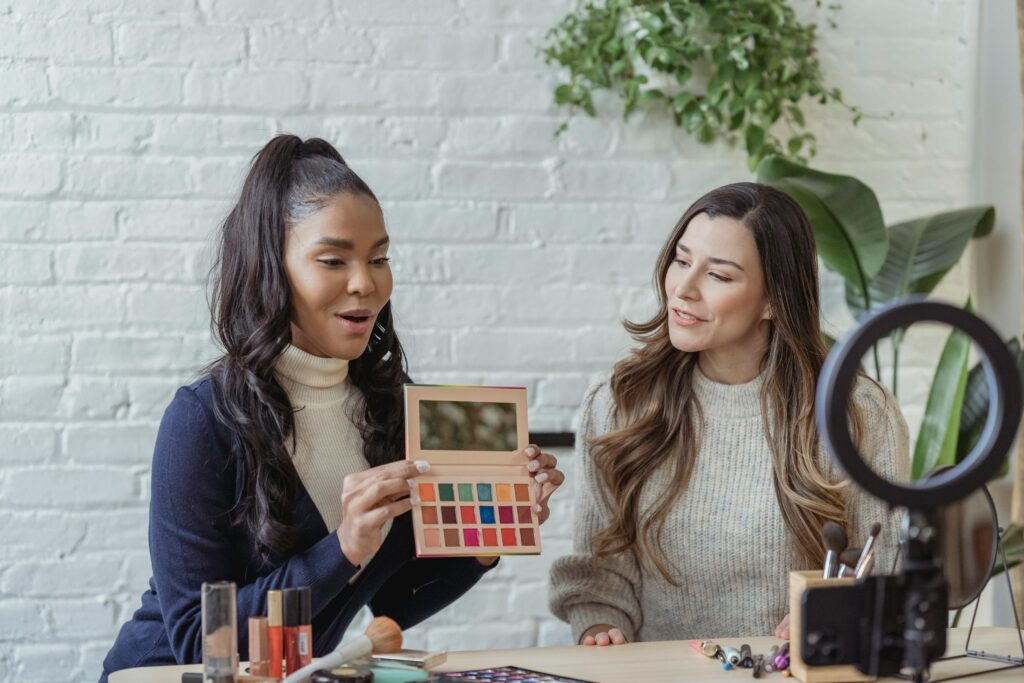 Two women sitting at a table with makeup products, one holding an eyeshadow palette, while a ring light and smartphone are set up in front of them.