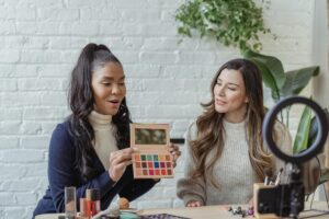 Two women sitting at a table with makeup products, one holding an eyeshadow palette, while a ring light and smartphone are set up in front of them.