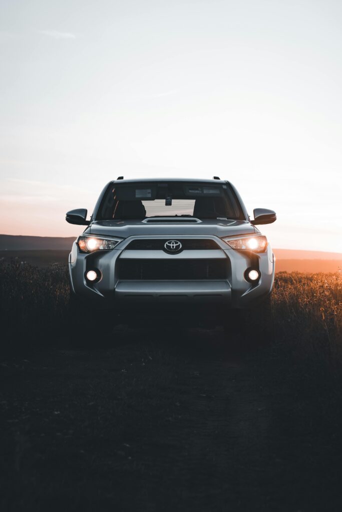 A silver SUV with its headlights on, set against a sunset sky.