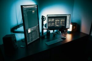 A dark home office setup with two monitors, one vertical and one horizontal, illuminated by ambient blue lighting, featuring speakers, a keyboard, mouse, and a small lamp.