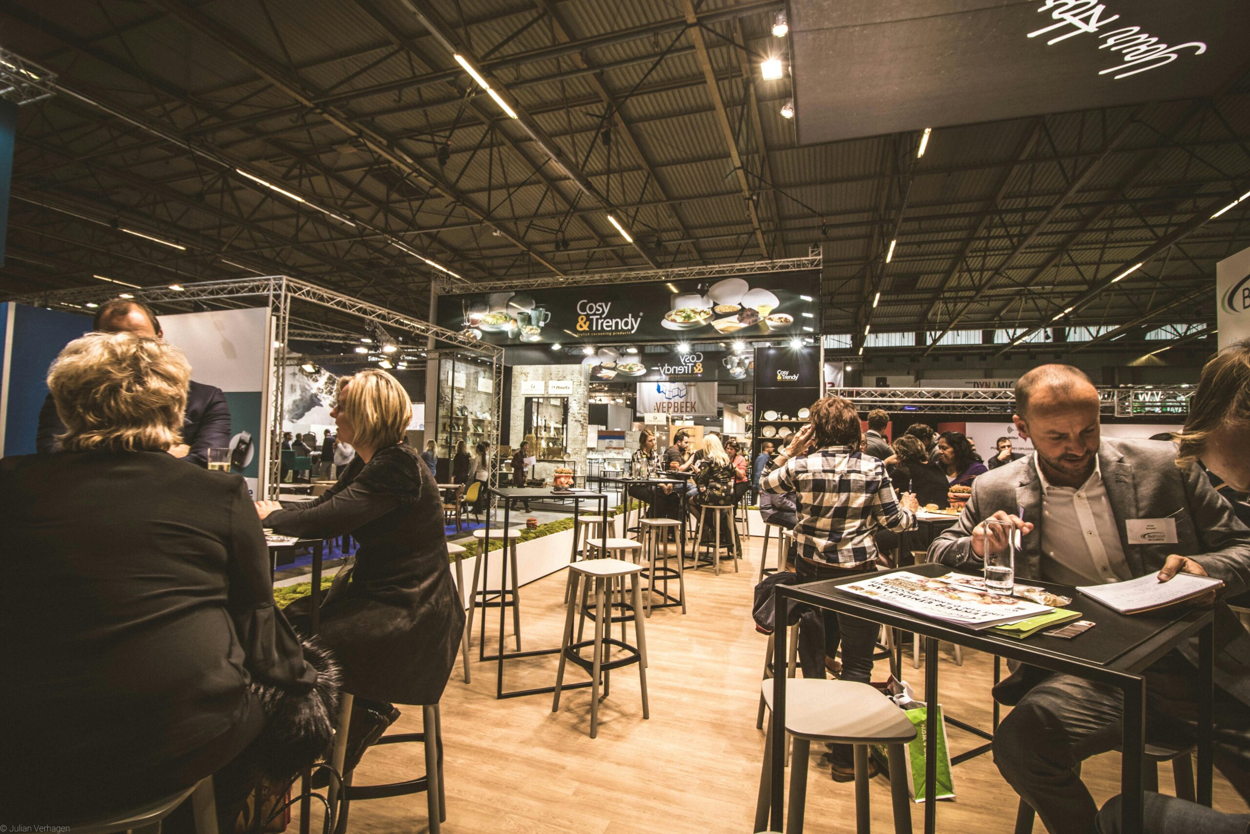 **Alt text:** People sitting and conversing at tables in a busy exhibition hall with a "Cosy & Trendy" banner visible in the background.