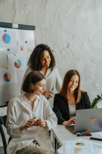 Three women smiling and working together at a desk with a laptop, in front of a whiteboard displaying charts.