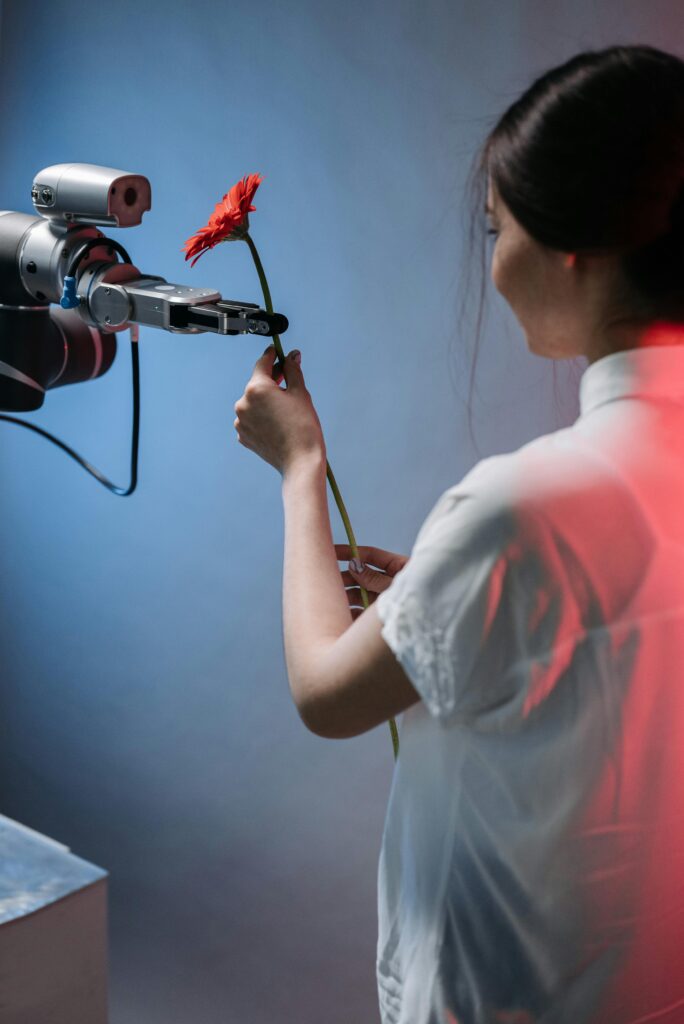 A woman in a white shirt is holding a red flower while a robotic arm reaches towards it.