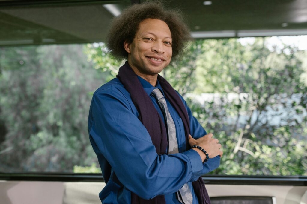 A person with curly hair smiles while standing in an office setting with a scenic view outside the window.