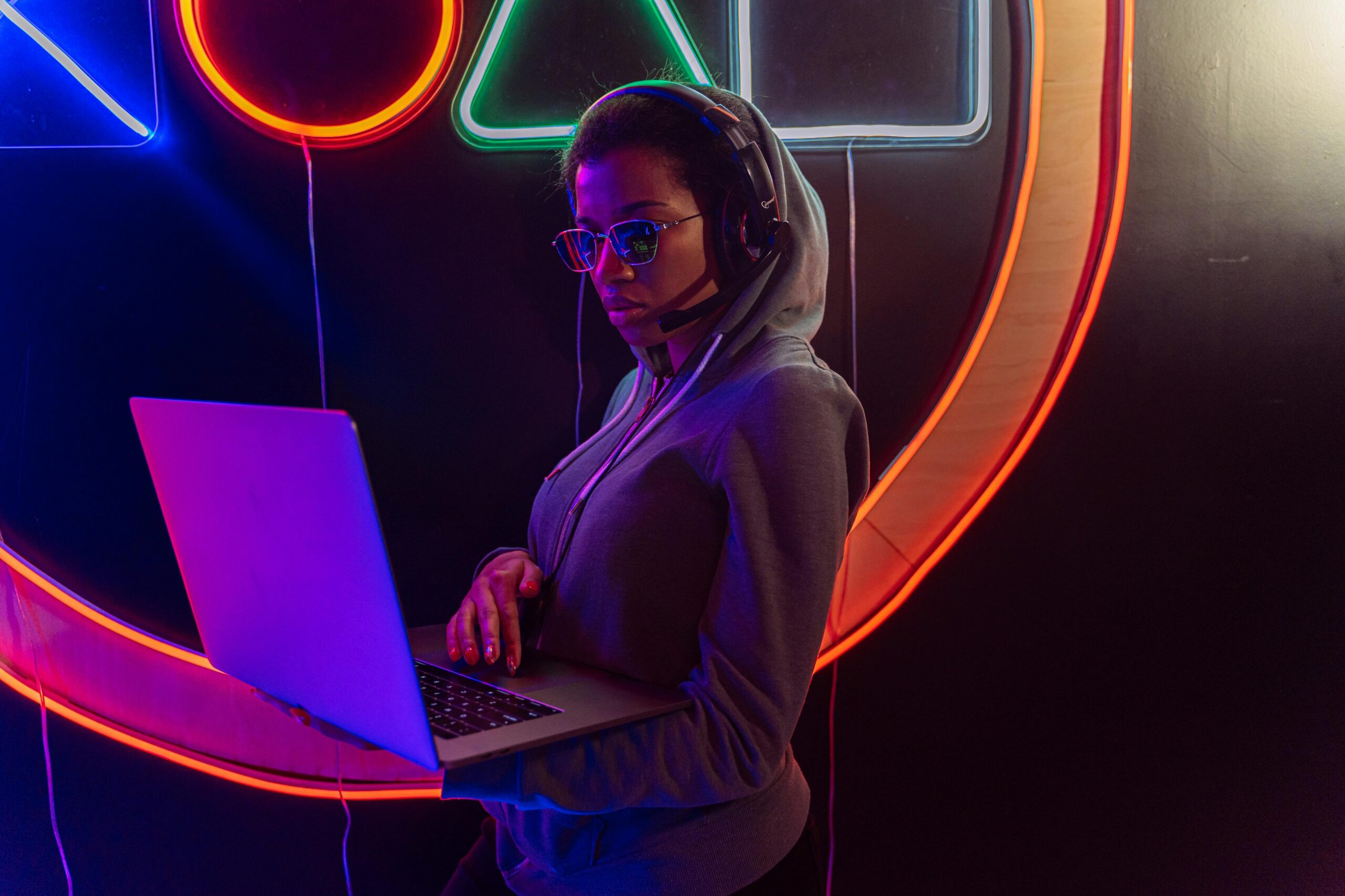 A person wearing sunglasses and a headset uses a laptop in front of a colorful neon light display.