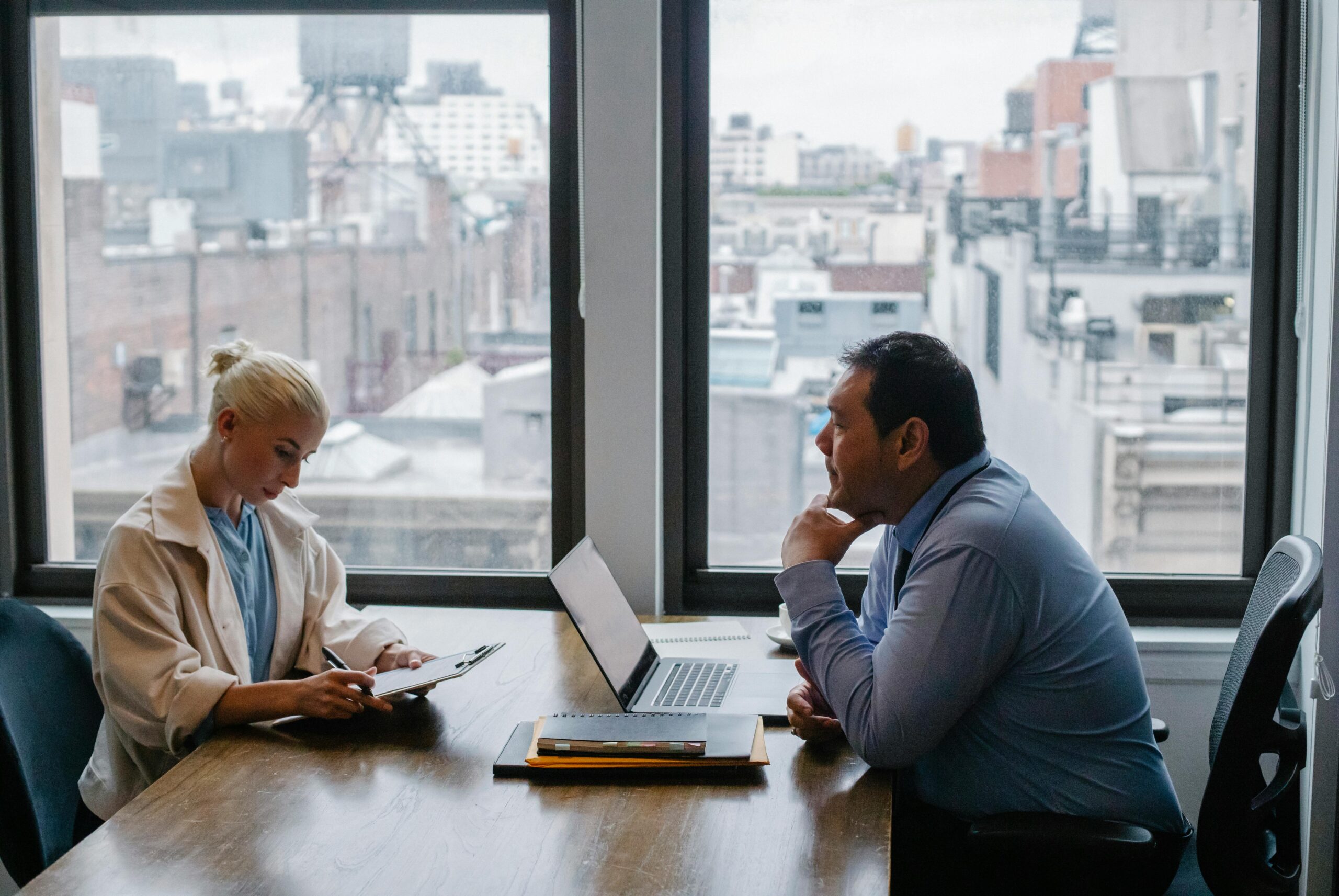 **Alt Text:** Two people are having a meeting in an office with large windows overlooking a cityscape; one is taking notes while the other listens attentively.
