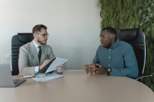 Two men having a discussion at a table in an office setting, one holding a clipboard and the other gesturing with his hands.