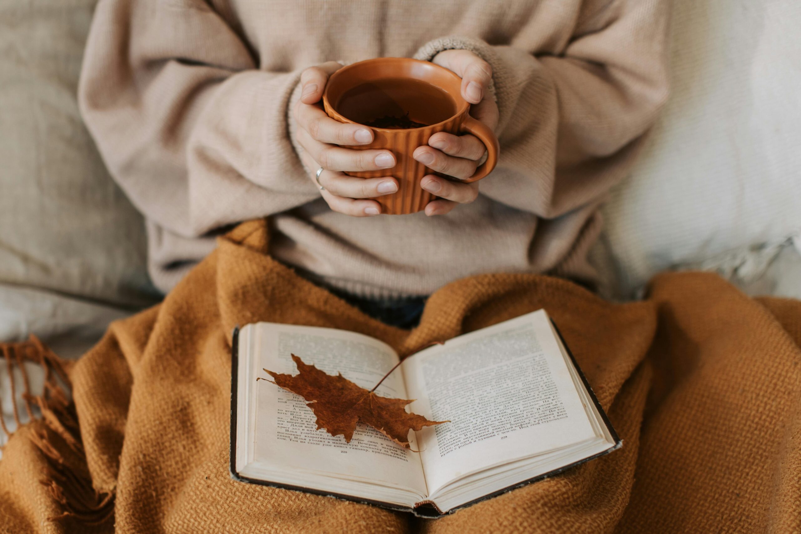 A person wearing a beige sweater holds a mug of tea above a book with a dry leaf resting on its pages, all atop a brown blanket.
