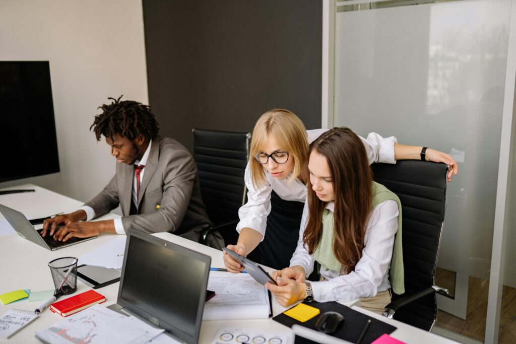 Three colleagues in a modern office, with two women focused on a tablet and a man working on a laptop.