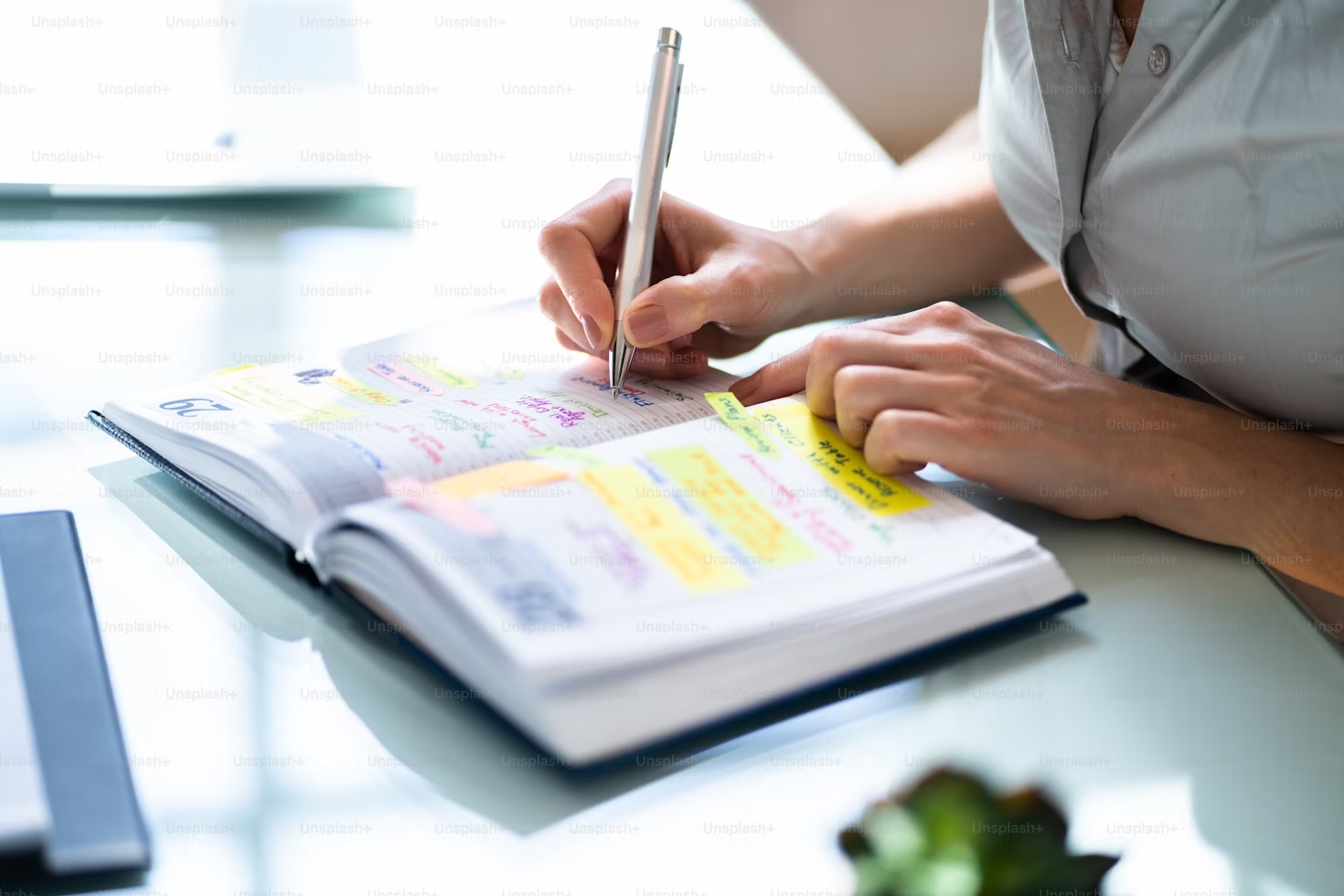 A person writing in a planner filled with colorful notes and sticky tabs on a glass desk.