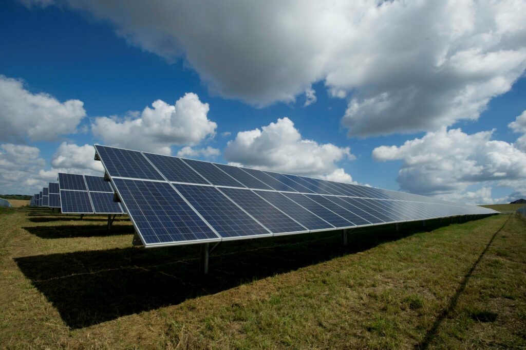 **Alt Text:** A row of solar panels on a grassy field under a partly cloudy blue sky.