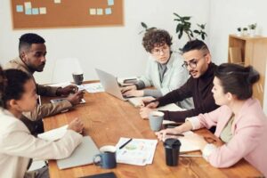 **Alt Text:** A group of five people sit around a wooden table in an office setting, engaged in a discussion with laptops and documents in front of them.