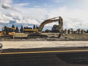 A large excavator is working on a construction site with piles of dirt and concrete cylinders nearby under a cloudy sky.