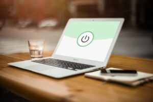 **Alt Text:** A laptop displaying a large power button icon on its screen is placed on a wooden table next to a glass of water, a smartphone, and a closed notebook.