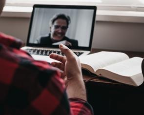 **Alt Text:** A person in a red plaid shirt is engaged in a video call on a laptop next to an open book.