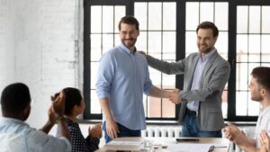 **Alt Text:** A man in a suit congratulates another man with a handshake in an office setting, as three colleagues seated at a table applaud.