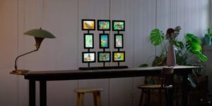 A wooden table with a green desk lamp on the left, a multi-photo frame with colorful images at the center, and a vase with flowers on the right; two stools are placed beside the table, with green plants visible in the background against a white paneled wall.