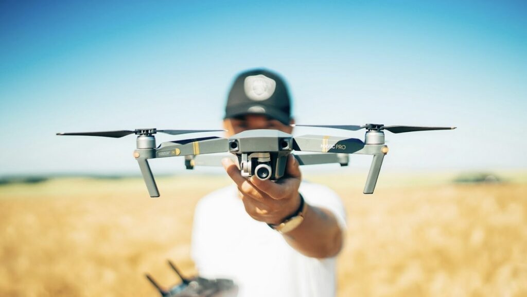 **Alt text:** A person holding a drone at arm's length in an open field with a clear blue sky in the background.