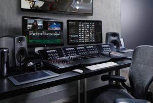 A professional video editing workstation with multiple monitors, speakers, editing console, and control panels on a desk.