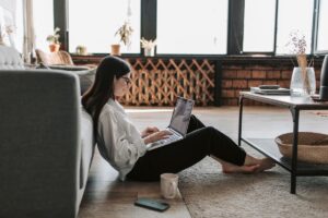 A woman sitting on the floor in a living room, using a laptop with a mug and phone nearby.