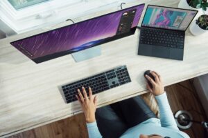 A person working at a desk with a wide monitor displaying a purple starry background and a laptop showing a photo editor.