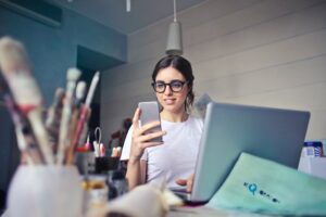**Alt text:** A woman wearing glasses is looking at her smartphone while working on a laptop at a desk with art supplies.
