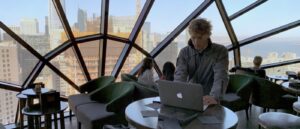 "A person working on a laptop in an upscale cafe with a panoramic city view through large windows."