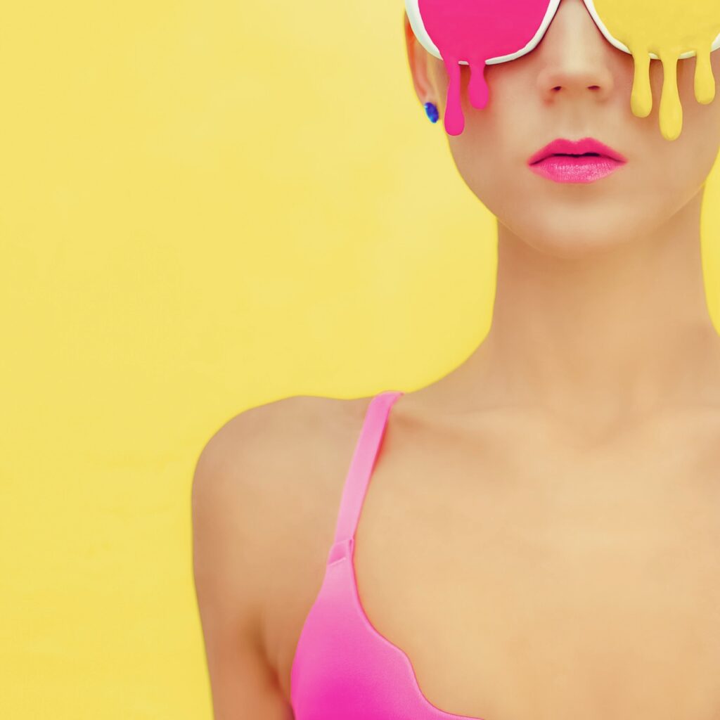 **Alt text:** A person wearing vibrant pink and yellow paint-drip style sunglasses and a pink top against a solid yellow background.