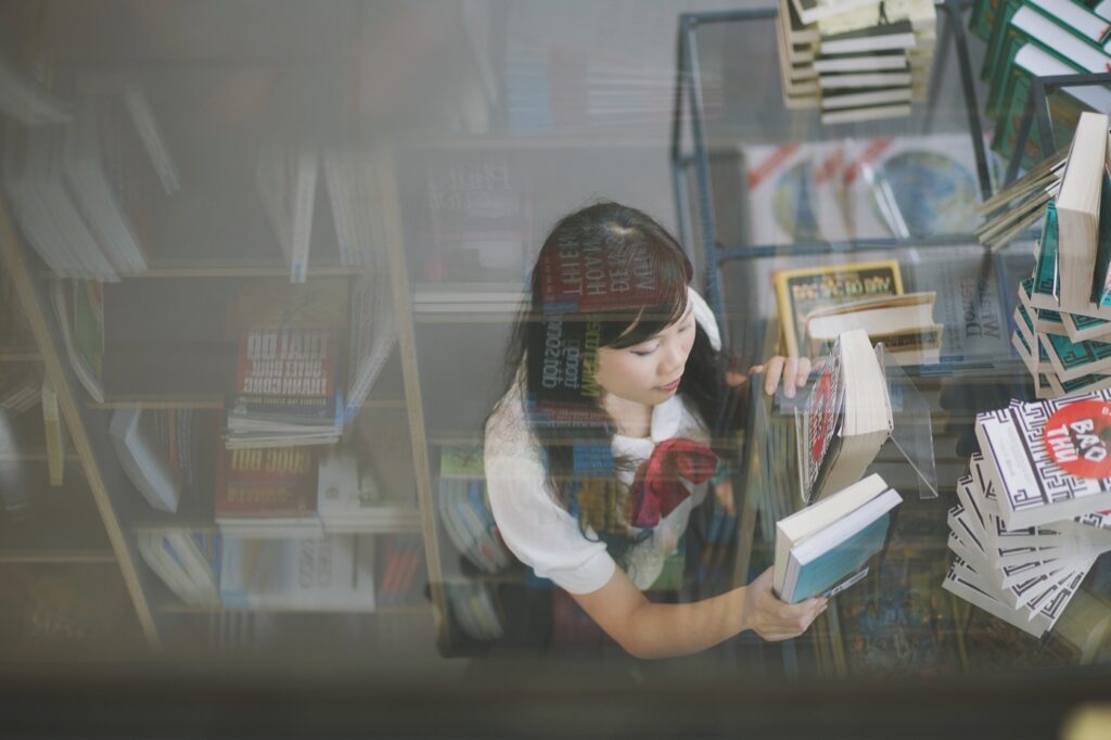 A young woman browses through books on a display table in a store, viewed from above through a glass surface.
