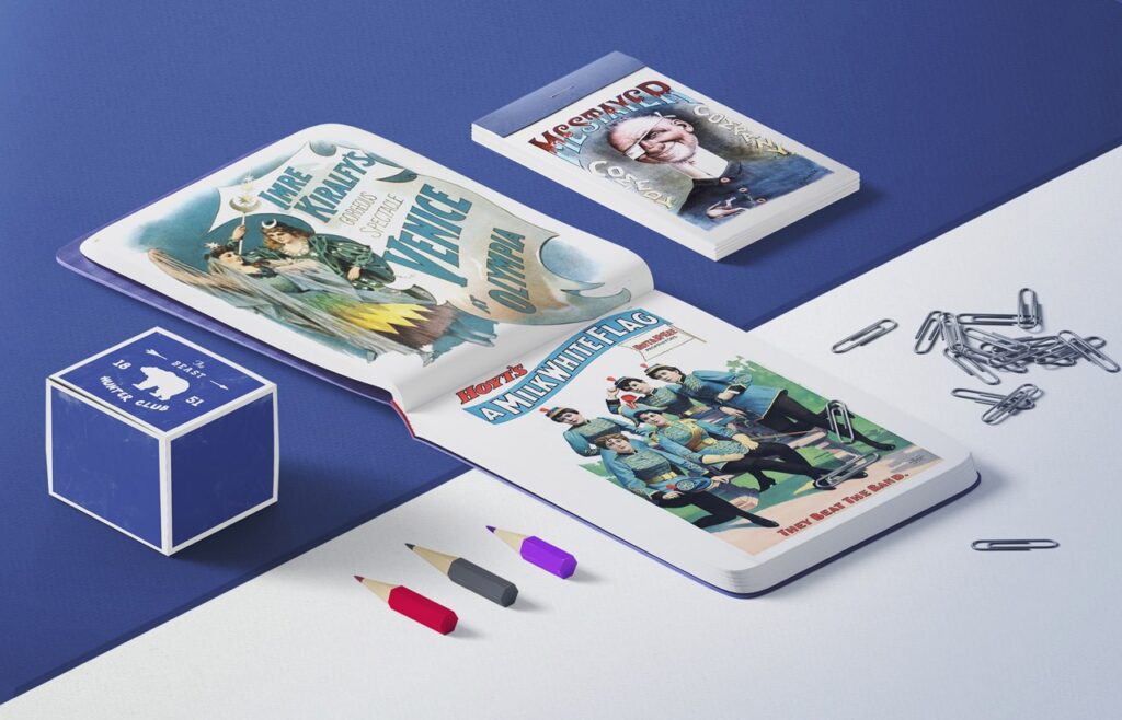 **Alt text:** An open book and a closed book with colorful vintage style illustrations, a cube with text, three pencils, and scattered paperclips on a blue and white surface.