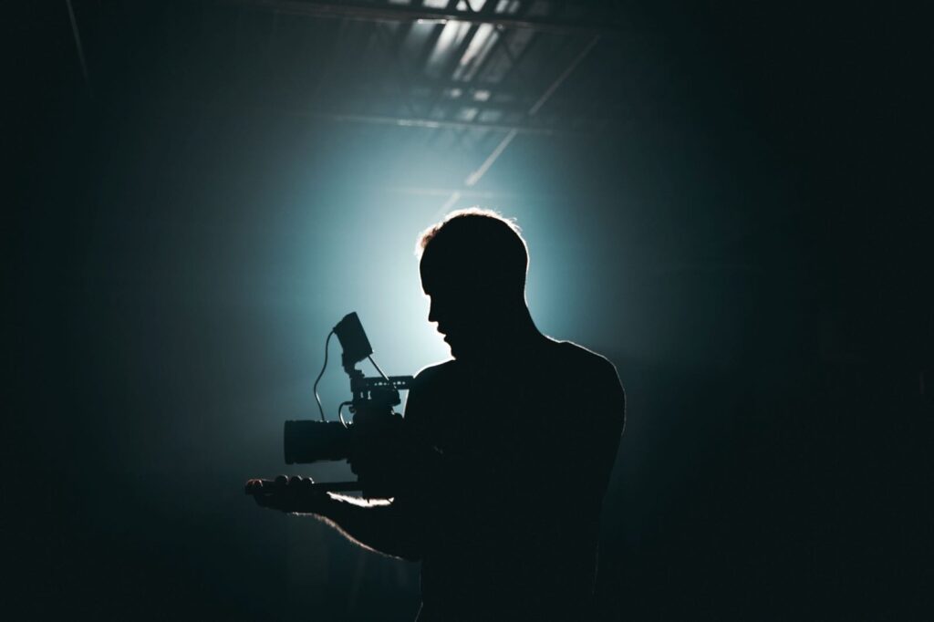 **Alt text:** A silhouette of a person holding a camera in a dimly lit setting with blue light in the background.
