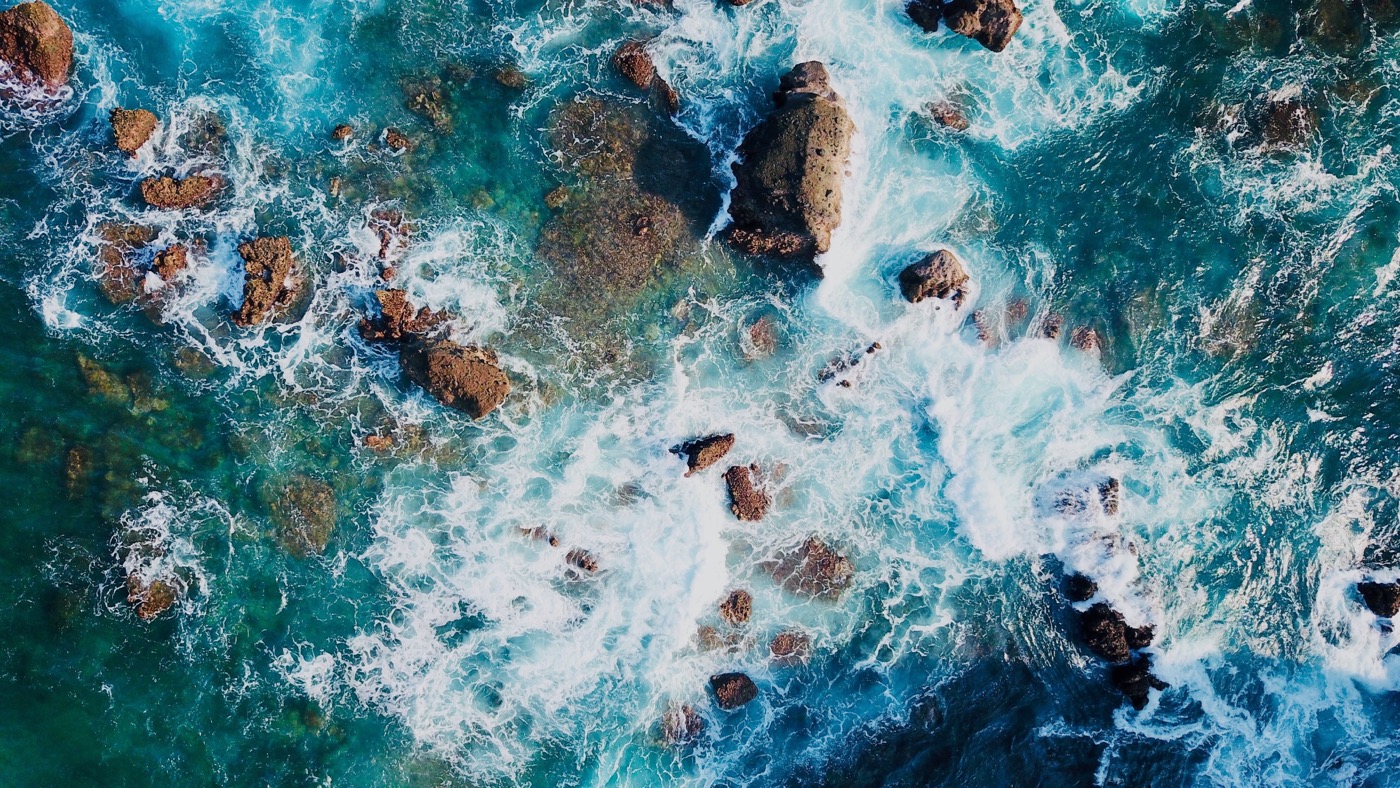 **Alt text:** Aerial view of turquoise ocean waves crashing over scattered rocks creating white foam.