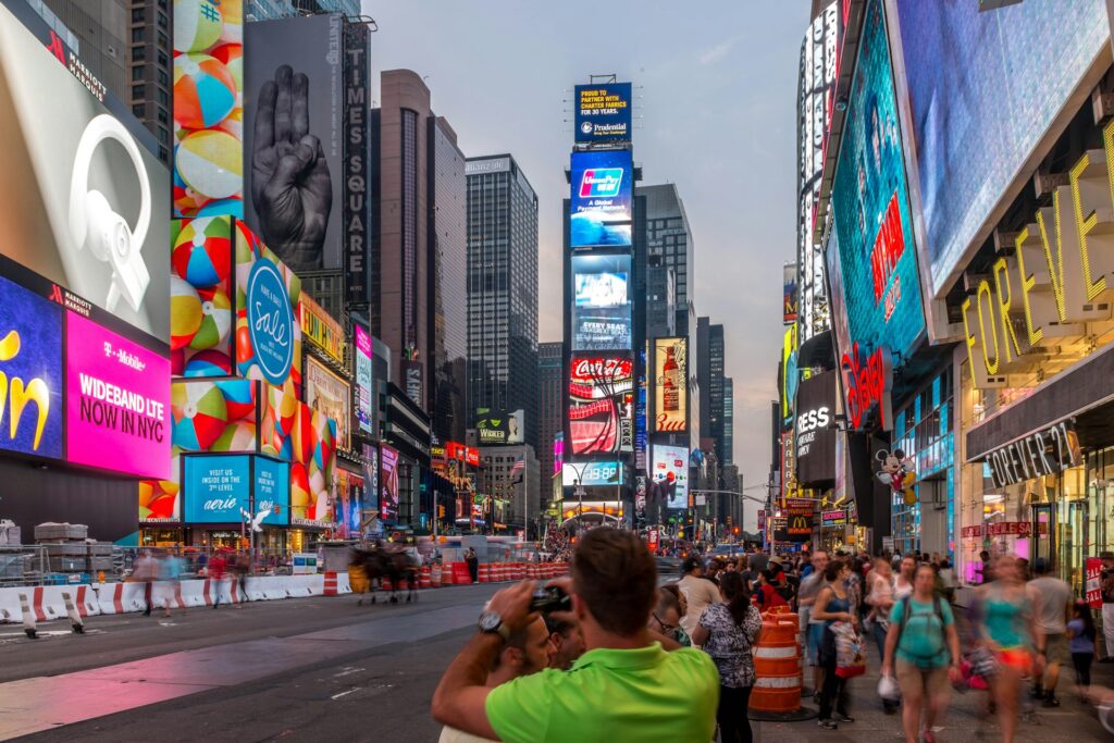 A busy evening scene in Times Square, New York City, featuring brightly lit billboards and crowds of people.
