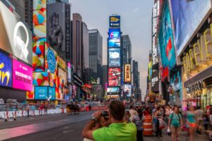 A busy evening scene in Times Square, New York City, featuring brightly lit billboards and crowds of people.