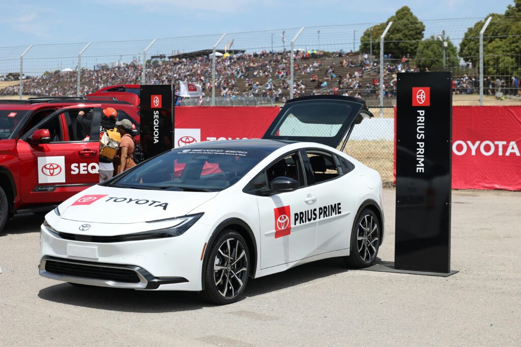 A white Toyota Prius Prime with its trunk open is displayed in front of a crowd at an outdoor event, alongside a red Toyota Sequoia.