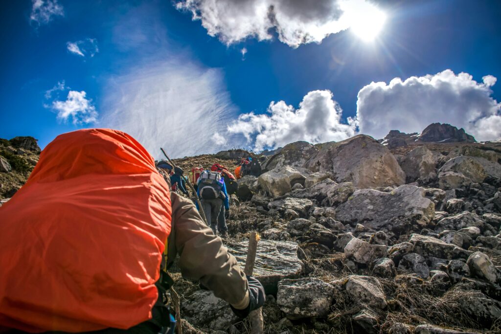 **Alt Text:** A group of hikers with backpacks climb a rocky mountain slope under a bright sun and blue sky.