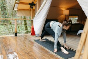 A woman practices yoga on a mat in a glamping tent surrounded by a forest setting.