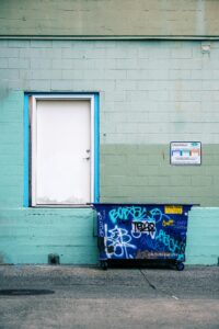 A white door with blue trim is set in a teal and green painted brick wall, beside a graffiti-covered blue dumpster.